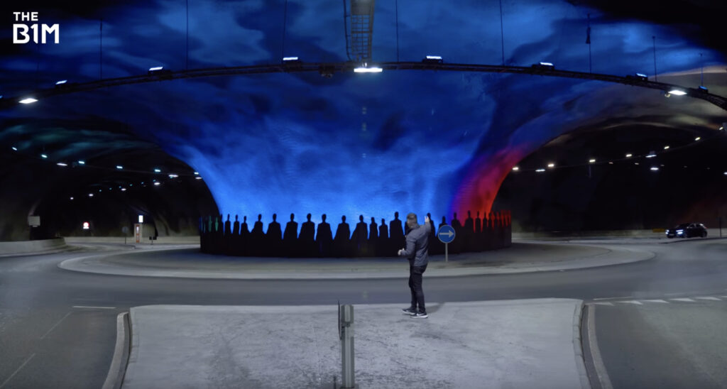 An underground roundabout. A man points down towards one tunnel exit while a car pulls into the roundabout.
The centre of the roundabout is lit up in soft blue and red colours with silhouettes of people visible.